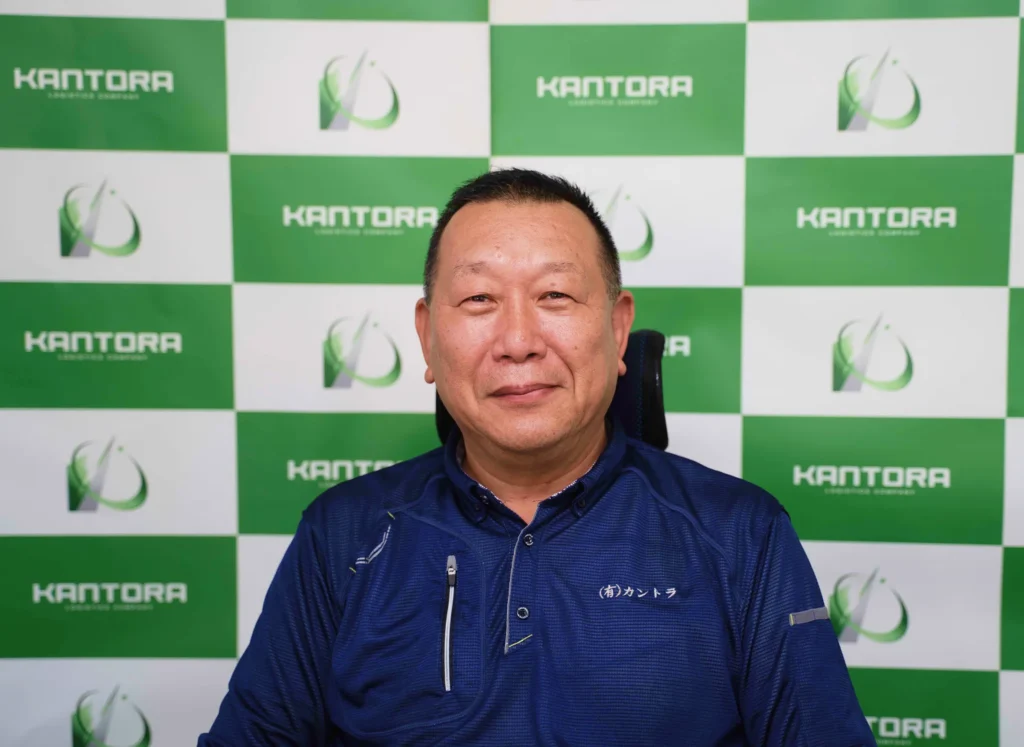 緑白のロゴ壁を背景に微笑む社長