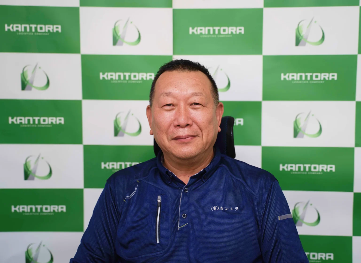 緑白のロゴ壁を背景に微笑む社長