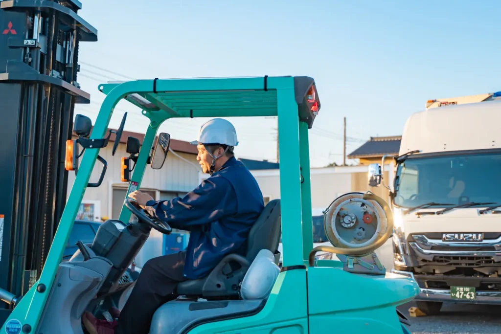 青緑色のフォークリフトを運転する白いヘルメットの男性